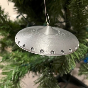 May include: A silver-colored UFO-shaped Christmas ornament with a string for hanging. The ornament has a textured surface and several small holes around the edge.