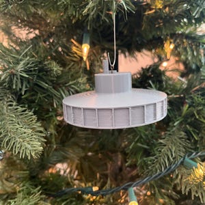 Può includere: Un ornamento a forma di UFO, stampato in 3D, di colore grigio, appeso a un albero di Natale. L'ornamento ha una base rotonda e piatta e una parte superiore a forma di cupola.