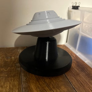 May include: A grey and black 3D printed model of a flying saucer on a stand. The saucer has a domed top with a series of small rectangular windows.