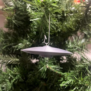 May include: A gray, saucer-shaped Christmas ornament, suspended by a silver hook, hangs against a backdrop of green Christmas tree branches. The ornament has a classic flying saucer design, perfect for a space-themed holiday.