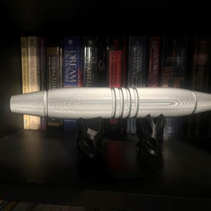 Könnte beinhalten: Ein silberfarbenes, zylindrisches Objekt mit drei Bändern, das auf einem schwarzen, dekorativen Ständer ruht. Das Objekt hat eine strukturierte Oberfläche und wird vor dem Hintergrund von Büchern in einem Regal ausgestellt.