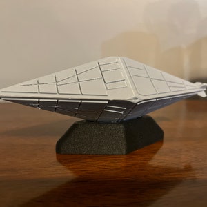 Puede incluir: Un modelo blanco de nave espacial de forma geométrica sobre una base negra. El modelo tiene una superficie detallada en forma de cuadrícula.