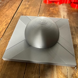 Puede incluir: Una base cuadrada gris, impresa en 3D, con un objeto en forma de cúpula gris, impreso en 3D, en la parte superior.