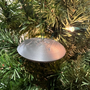 Pode incluir: Um enfeite de Natal redondo em forma de disco voador de cor prateada pendurado em uma árvore de Natal artificial verde.