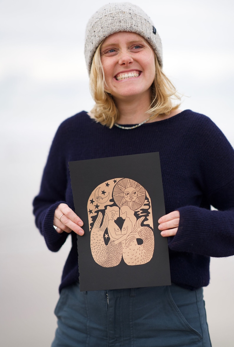 K&ouml;nnte beinhalten: Eine Person h&auml;lt einen schwarzen Druck mit einer kupferfarbenen Illustration von zwei Meerjungfrauen, die sich unter einem Halbmond und der Sonne umarmen. Die Meerjungfrauen haben langes, flie&szlig;endes Haar und Schw&auml;nze mit Schuppen. Der Hintergrund ist mit Sternen gef&uuml;llt.