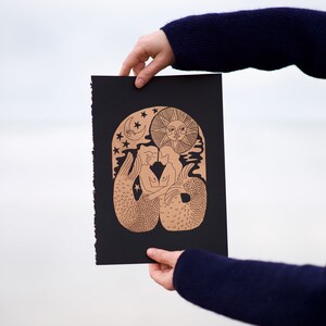 K&ouml;nnte beinhalten: Ein schwarzer Druck mit einem goldenen Design von zwei sich umarmenden Meerjungfrauen, umgeben von Sternen, einem Halbmond und einer Sonne. Der Druck wird von einer Hand gehalten.