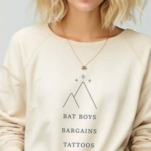 Puede incluir: Una sudadera de color crema con un gráfico de montaña negro y el texto "BAT BOYS BARGAINS TATTOOS". La modelo lleva un collar de oro.