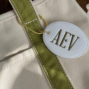 Puede incluir: Primer plano de un bolso tote de lona con una correa verde y una etiqueta ovalada blanca. La etiqueta presenta las letras bordadas "AEV" en hilo verde a juego. Un anillo metálico dorado conecta la etiqueta a la correa.