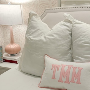 May include: A bedroom scene featuring a white bed with large, fluffy pillows. A decorative pillow with the letters "TMM" in pink is on the bed. A pink lamp sits on a white nightstand with books. The wallpaper has a pink floral pattern.
