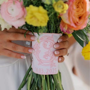 Puede incluir: Un colorido ramo de flores con flores rosas, amarillas y naranjas. Los tallos están envueltos con una tela rosa adornada con un diseño decorativo y la fecha 10.03.2025.