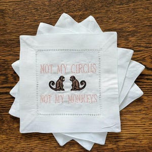 May include: White square cocktail napkins with a decorative stitched border. The napkins feature the embroidered phrase "NOT MY CIRCUS NOT MY MONKEYS" in light pink, along with two monkey illustrations. The napkins are stacked on a wooden surface.