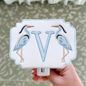 May include: A decorative night light featuring a white rectangular design with a light blue border. The design includes a light blue letter "V" and two embroidered blue herons. The night light is held by a person.