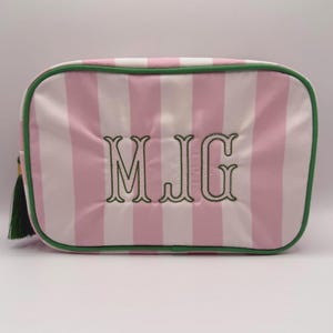 Puede incluir: Bolsa de cosméticos a rayas rosas y blancas con ribete verde y borla verde. La bolsa presenta las iniciales bordadas "MJG" en verde. La bolsa es rectangular con esquinas redondeadas.