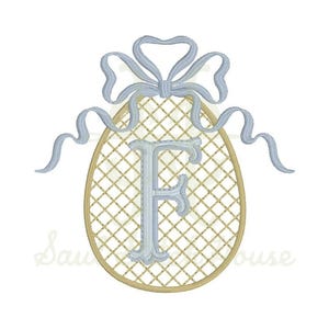 Puede incluir: Un diseño de bordado que presenta una letra F azul claro centrada en un óvalo dorado con un patrón de rejilla entrecruzada. Un lazo de cinta azul claro con extremos ondulados se coloca en la parte superior del óvalo, completando el motivo.