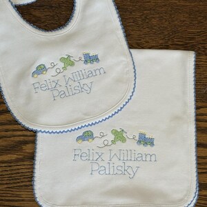 May include: White baby bib and burp cloth set with blue trim. Embroidered with a car, aeroplane, and train design, and the name "Felix William Palisky" in blue thread. The bib has a snap closure.