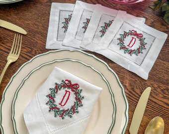 May include: White linen napkins with a green and red embroidered wreath design and the letter "D". The napkins are placed on cream-colored plates with a green trim, alongside gold cutlery, creating a festive table setting.