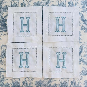 Könnte beinhalten: Vier weiße Leinenuntersetzer mit einem türkis gestickten Buchstaben "H" in der Mitte. Jeder Untersetzer hat eine dekorative, genähte Bordüre. Die Untersetzer liegen auf einer blau-weiß gemusterten Oberfläche.