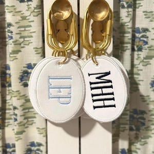May include: White oval keychains with embroidered initials hang from gold-colored hardware. One keychain has the initials "LEP" in light blue, and the other has "MHH" in black. The background features floral patterned fabric.