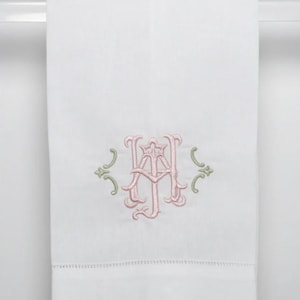 May include: A white hand towel featuring a central embroidered monogram in light pink thread, flanked by small sage green decorative flourishes. The towel has a hemstitched border detail near the bottom edge and a smooth, plain fabric texture.