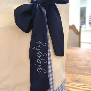 May include: A cream tote bag with blue gingham detailing and handles. A navy blue ribbon is tied in a bow, with the word "giggley" embroidered in light blue script on the ribbon.