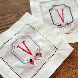 May include: Two square, off-white linen coasters with embroidered details. Each coaster features a shield design with a red letter "V" and a blue border. Small American flags are embroidered below the shield. The edges have a decorative stitched border.