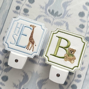 May include: Two decorative night lights with embroidered designs. One features the letters "HSE" and a giraffe, the other has the letter "B" and a teddy bear. Both have a white base and are set against a patterned background.