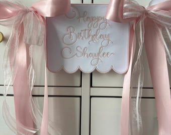 May include: A white sign with the text "Happy Birthday Shaylee" embroidered in pink script. The sign is adorned with pink satin bows and sheer ribbons, creating a festive and celebratory look. The sign is hanging on a white cabinet.