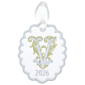 May include: A white scalloped ornament featuring an embroidered light green and light blue letter V in a decorative font. The year 2026 is embroidered in light blue at the bottom. A white satin ribbon loop is attached to the top for hanging.