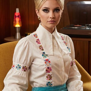 May include: A long-sleeved white button-down shirt featuring vertical panels of colorful floral embroidery along the front and sleeves, paired with a solid turquoise blue mini skirt. The shirt has a pointed collar and gathered sleeve cuffs.