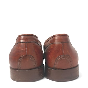 May include: A pair of brown leather loafers shown from the back, featuring a smooth finish with visible natural grain, reinforced heel stitching, and dark brown stacked heels. The leather displays subtle variations in tone and light surface scuffs.