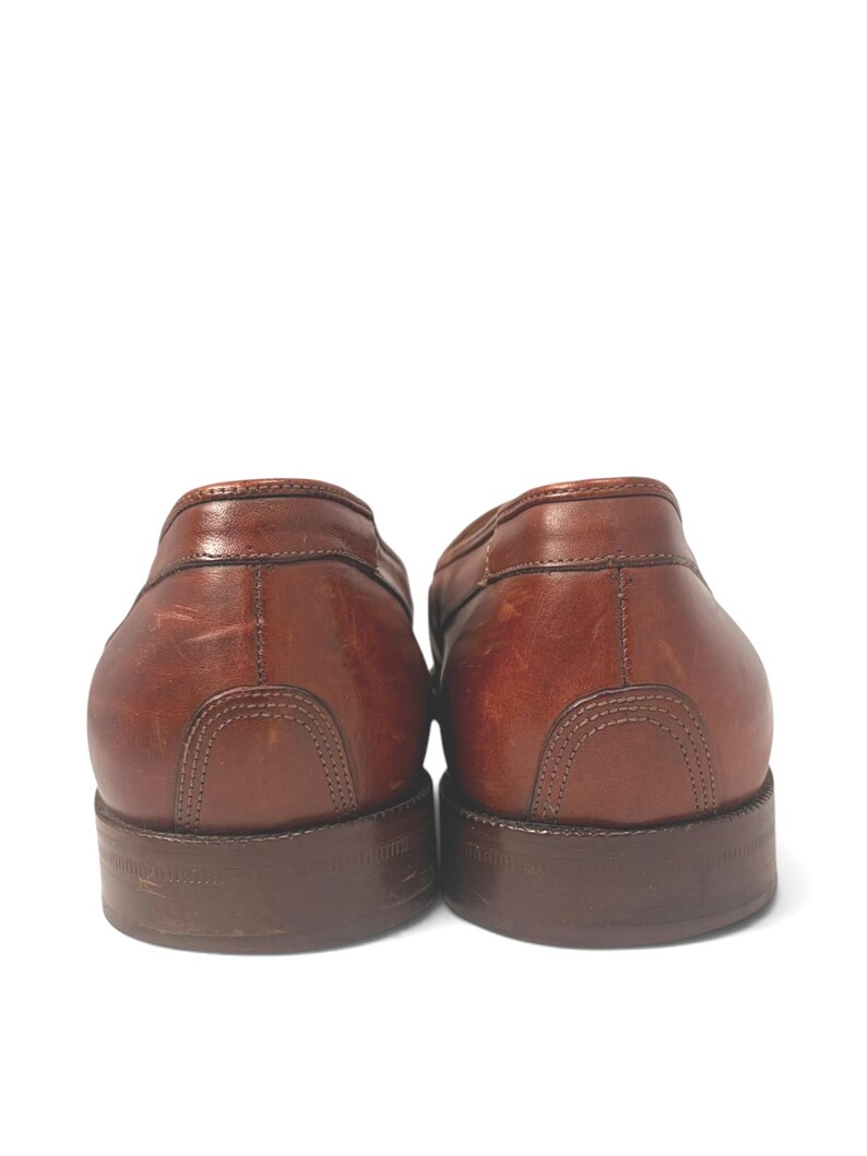 May include: A pair of brown leather loafers shown from the back, featuring a smooth finish with visible natural grain, reinforced heel stitching, and dark brown stacked heels. The leather displays subtle variations in tone and light surface scuffs.
