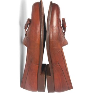 May include: A pair of brown leather loafers featuring a tassel detail on the top of each shoe. The shoes have a rounded toe, a low stacked heel, and visible stitching along the vamp. The leather has a smooth, polished finish with natural grain.