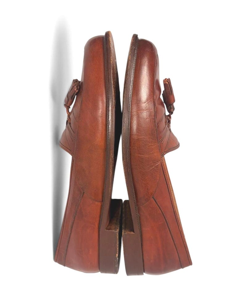 May include: A pair of brown leather loafers featuring a tassel detail on the top of each shoe. The shoes have a rounded toe, a low stacked heel, and visible stitching along the vamp. The leather has a smooth, polished finish with natural grain.