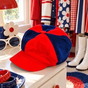 May include: A corduroy newsboy cap featuring alternating panels of vibrant red and deep navy blue fabric. The hat has a structured, rounded crown and a stiff, matching red visor, showcasing a classic multi-panel construction.
