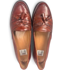 May include: A pair of brown leather tassel loafers featuring a moccasin-style stitched toe, a decorative leather fringe, and a small tassel detail on the vamp. The interior soles are lined with tan leather and feature a Cable & Co. brand label.