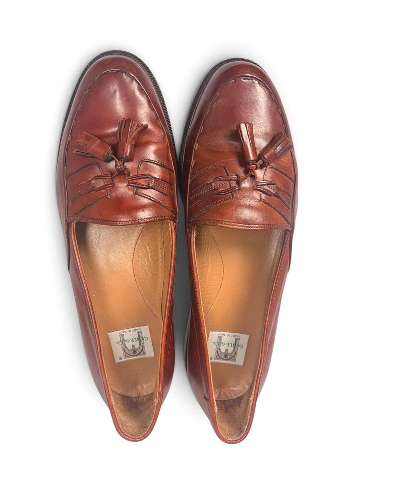 May include: A pair of brown leather tassel loafers featuring a moccasin-style stitched toe, a decorative leather fringe, and a small tassel detail on the vamp. The interior soles are lined with tan leather and feature a Cable & Co. brand label.