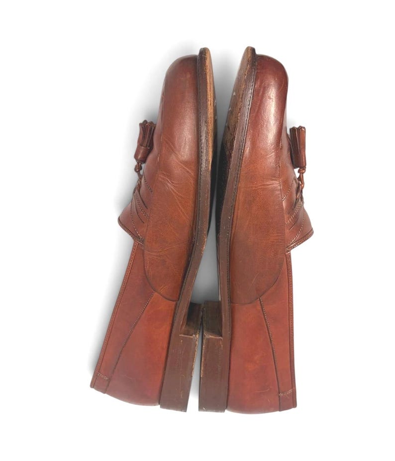 May include: A pair of brown leather tassel loafers featuring a rounded toe, a low stacked heel, and decorative stitching along the vamp. The shoes have a smooth finish with visible natural creasing on the upper surface and a dark brown sole.