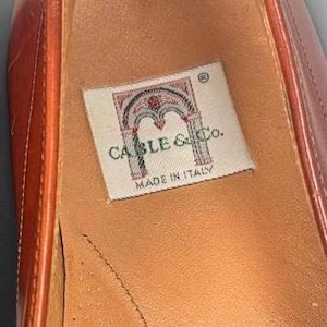 May include: A tan leather shoe interior featuring a rectangular white fabric label with a red and grey archway graphic. The text on the label reads CABLE & Co. and MADE IN ITALY. The shoe has visible stitching along the side and a smooth finish.