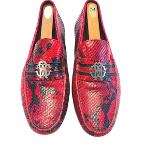 May include: Pair of red and black snakeskin-patterned loafers. Each shoe features a silver-tone emblem and a black band across the vamp. Wooden shoe trees are inserted inside the shoes.