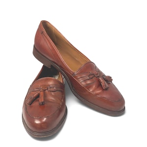 May include: A pair of brown leather loafers featuring a classic moccasin-style toe, decorative leather tassels on the vamp, and a low stacked heel. The shoes have a smooth finish with visible stitching along the apron and a tan interior lining.