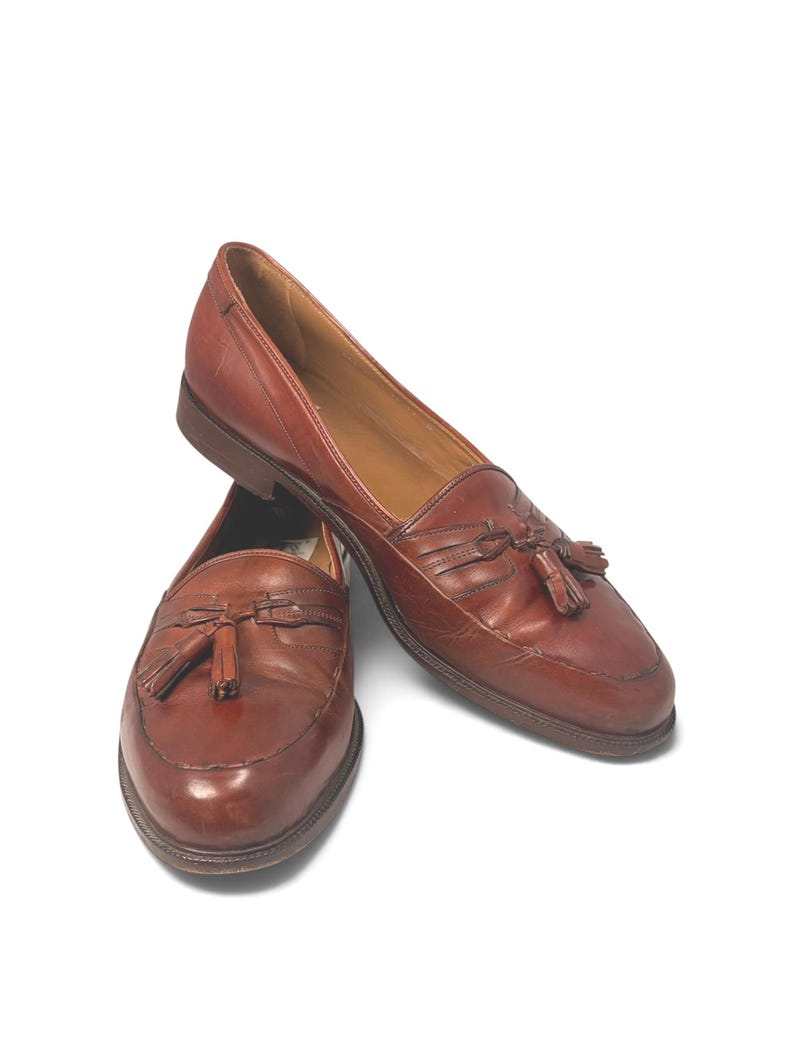 May include: A pair of brown leather loafers featuring a classic moccasin-style toe, decorative leather tassels on the vamp, and a low stacked heel. The shoes have a smooth finish with visible stitching along the apron and a tan interior lining.