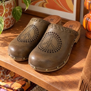 May include: A pair of olive green leather clogs featuring a perforated peace sign design on the vamp, a stitched leather trim along the collar, and a wooden sole secured with small brass-toned metal studs around the base of the shoe.
