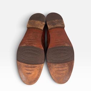 May include: A pair of brown leather shoes shown from the bottom view, featuring worn soles with dark brown textured rubber inserts at the midfoot and heel, and a stacked heel design with visible signs of wear and scuffing on the leather surface.