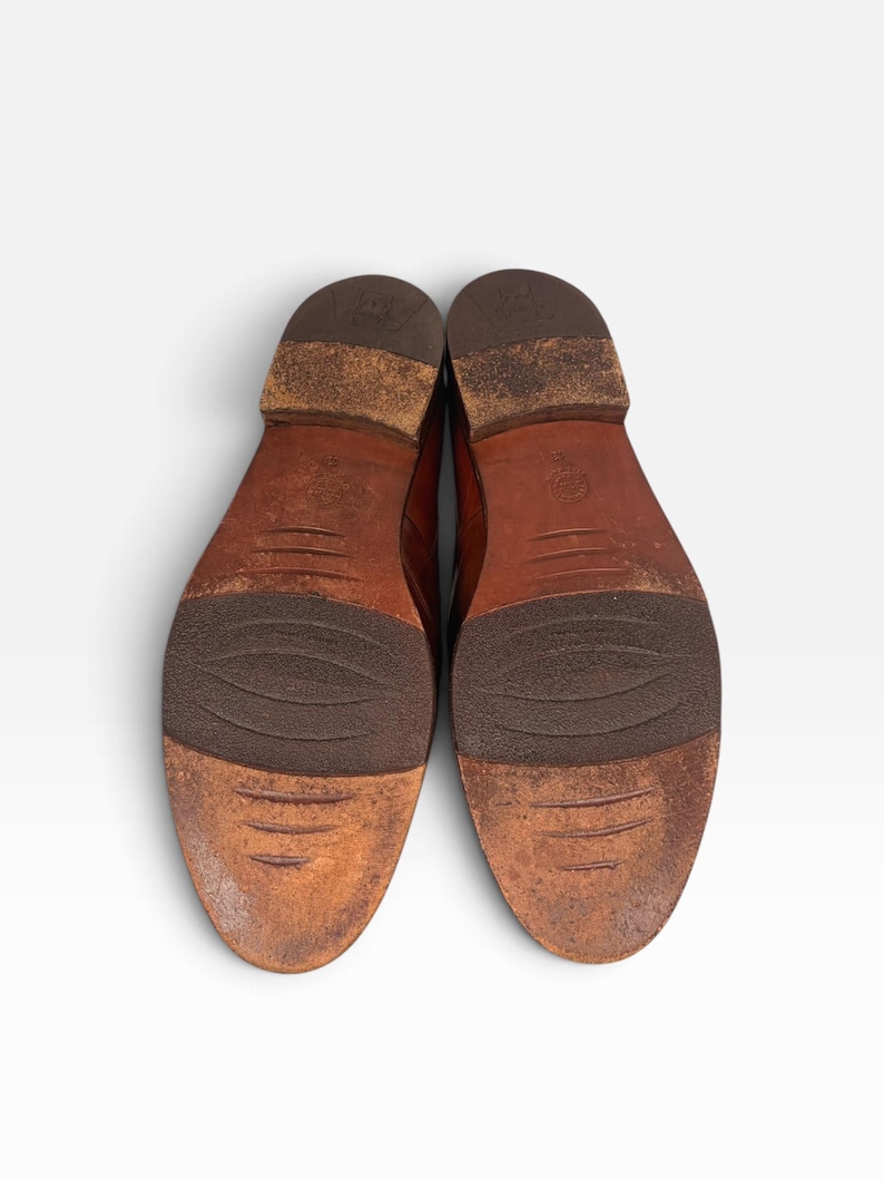 May include: A pair of brown leather shoes shown from the bottom view, featuring worn soles with dark brown textured rubber inserts at the midfoot and heel, and a stacked heel design with visible signs of wear and scuffing on the leather surface.
