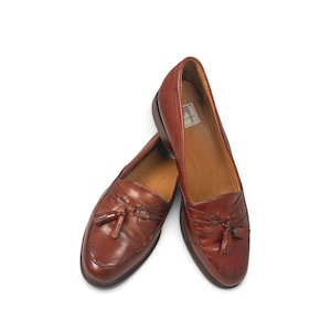 May include: A pair of brown leather loafers featuring a classic moccasin-style toe, decorative tassels on the vamp, and a low stacked heel. The interior of the shoe displays a white label with text, and the leather has a polished finish.