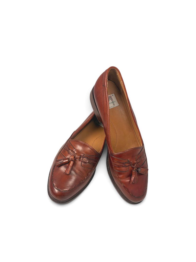 May include: A pair of brown leather loafers featuring a classic moccasin-style toe, decorative tassels on the vamp, and a low stacked heel. The interior of the shoe displays a white label with text, and the leather has a polished finish.