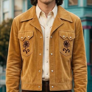 May include: Mustard yellow suede jacket with floral embroidery on the pockets. The jacket has a button-up front and a collared neckline. Worn over a cream-colored button-down shirt and brown pants.
