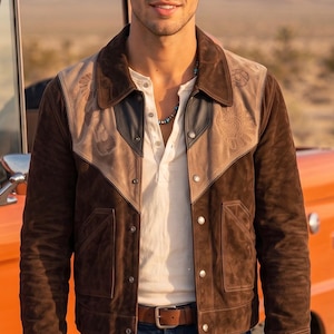 May include: A dark brown suede jacket featuring a lighter tan suede yoke with embroidered circular patterns, a pointed collar, silver-tone metal snap buttons, and two front pockets. The jacket is worn over a cream-colored henley shirt and blue jeans.