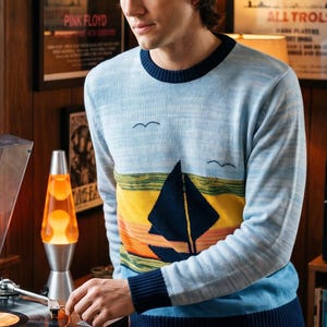 May include: A long-sleeve crew neck sweater featuring a light blue heathered knit base with a dark navy ribbed collar, cuffs, and hem. The chest displays a graphic of a dark navy sailboat on a yellow and orange horizon with two small birds.