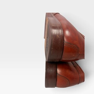 May include: A pair of stacked brown leather shoes featuring a smooth finish, visible stitching along the heel counter, and a dark brown stacked wooden heel. The shoes are positioned one above the other, showcasing the side profile and heel construction.